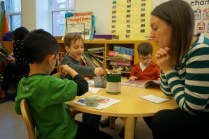 Teacher sits at a table with students, instructing them as they use watercolor paints.