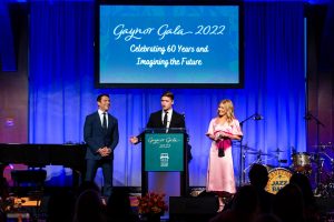 Joaquin Consuelos presents his speech to the Gaynor Gala attendees as his parents Mark Consuelos and Kelly Ripa stand on either side of him. Kelly is wearing a pink dress, while Mark and Joaquin are wearing suits.