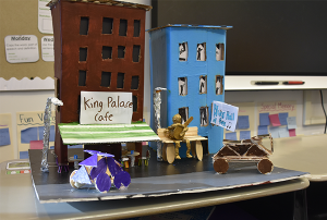 Class project of cardboard buildings; in front of the buildings are lamp posts made out of aluminum foil and cars made from wood pieces