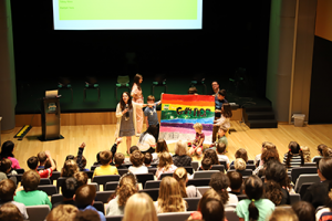 Students gathered in an assembly look at a group of students holding a Gaynor flag