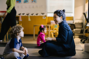 An Early Childhood teacher sits on the floor with a student.