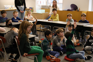 Students are sitting in a desks in a u-shape, and students sitting on the floor in the middle are raising their hands