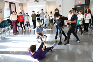 Blue Cluster students assembled with one student lying on the ground reaching up towards the other four. Another student's arms are being held by two students, one on each side of him, while the last student crouches on the ground,