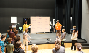 Two Teaching Artists stand on the stage in front of the students, on either side of a whiteboard